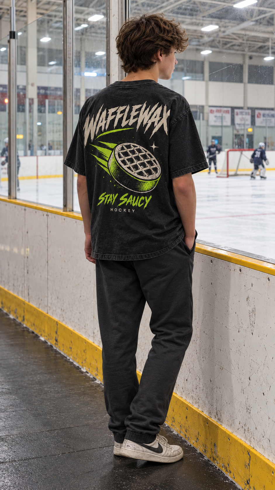 Back view of the WaffleWax Stay Saucy tee with cream and lime waffle-puck artwork, worn at an ice rink.